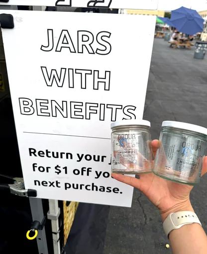 Two empty Obour jars held up next to the market stand's sign: Jars with benefits, return your jar for $1 off your next purchase