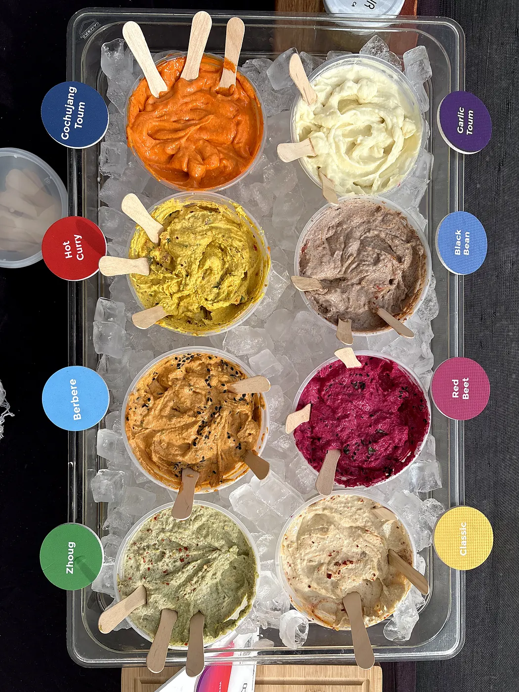A tray of hummus and toum samples on ice, each one labeled