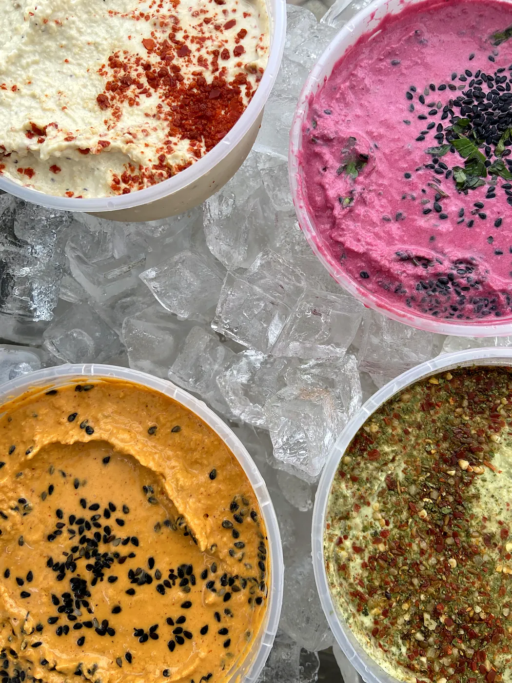 Four tubs of garnished hummus on ice, seen from above