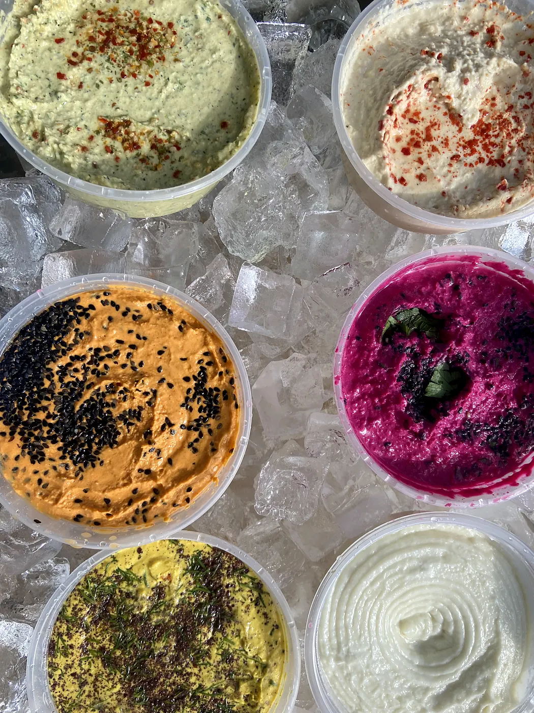 Six garnished tubs of hummus and toum on ice