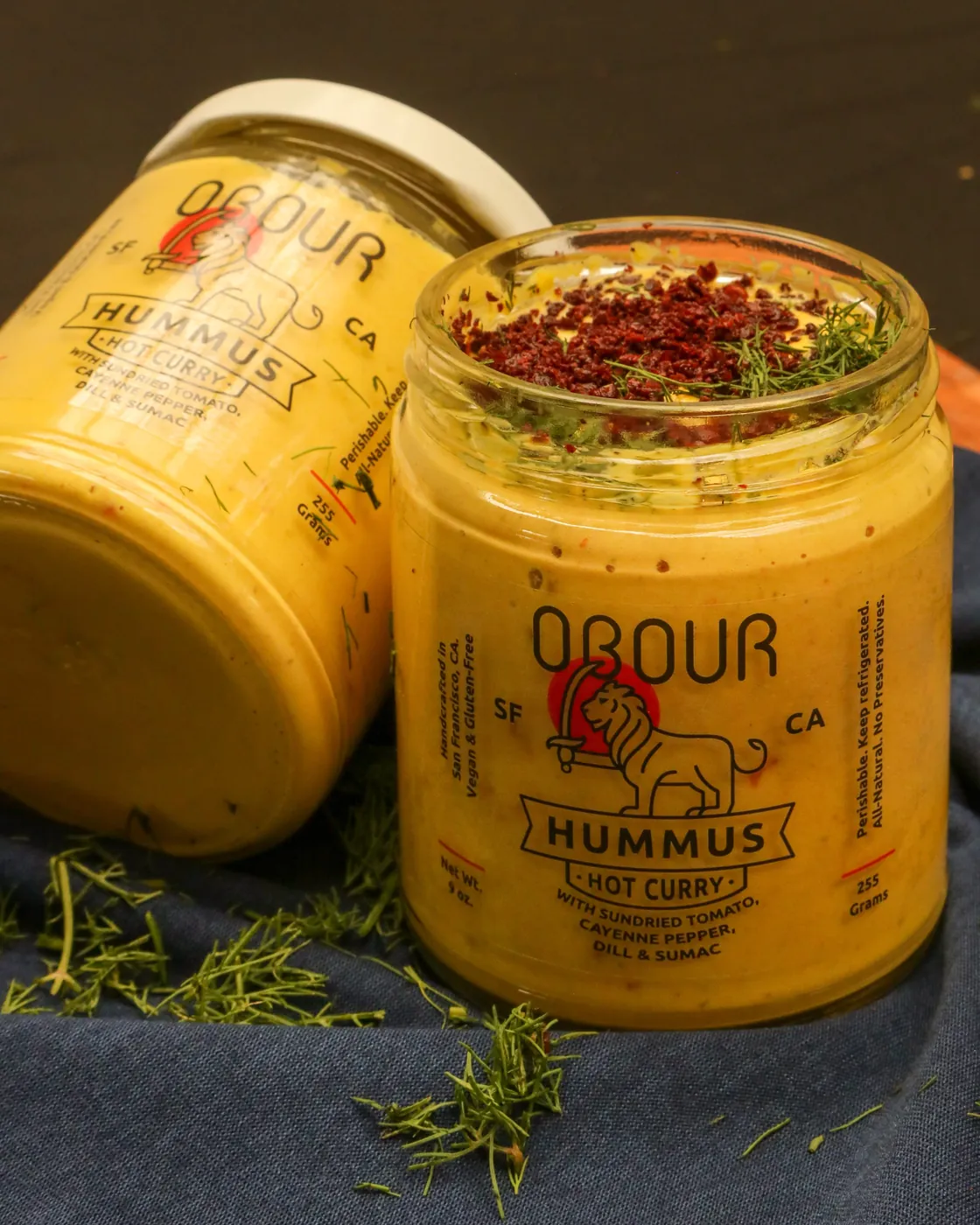 An open jar of Obour Hot Curry hummus topped with sumac and dill, next to a closed jar